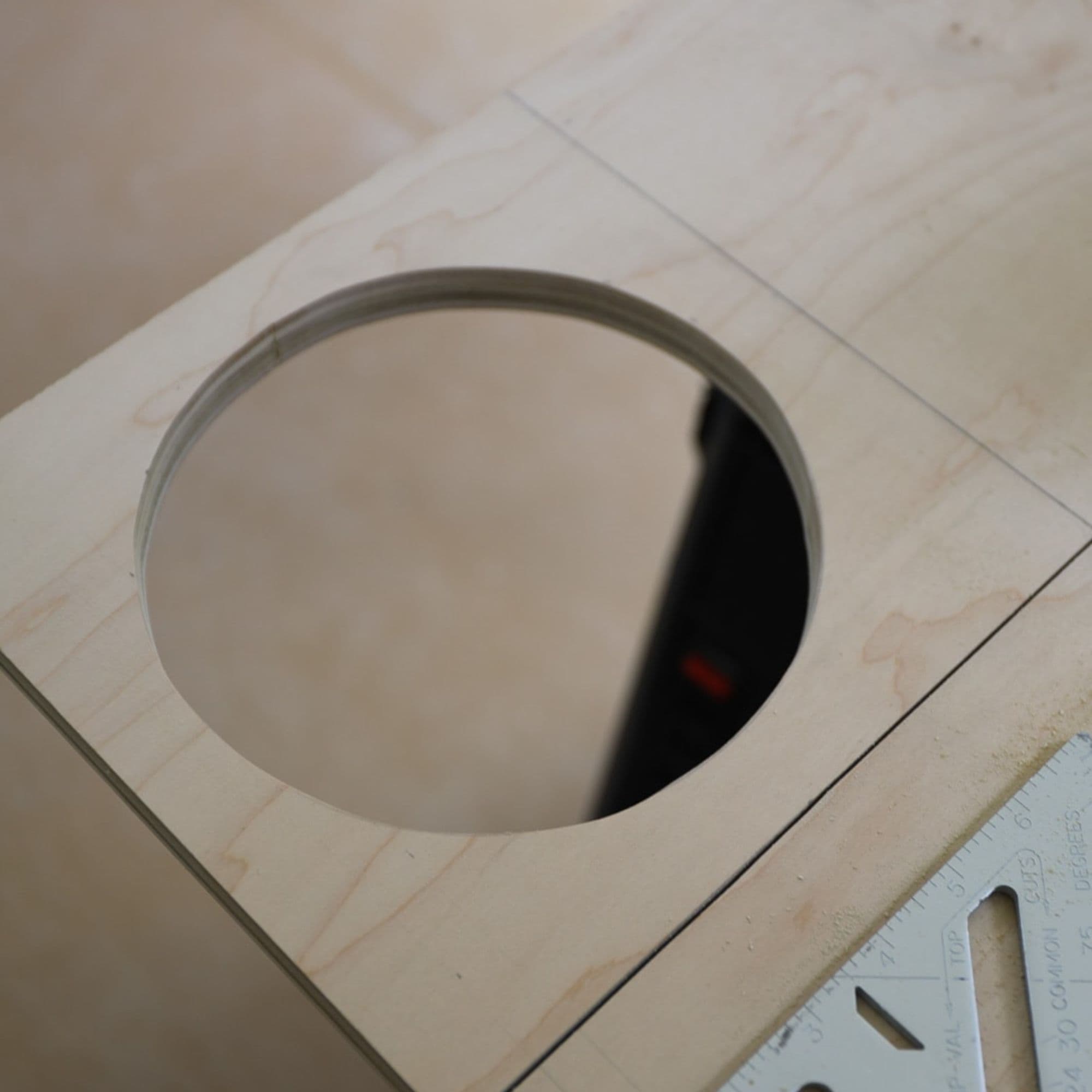 Circular opening cut into a sheet of plywood, with pencil guide lines and a measuring square visible nearby.