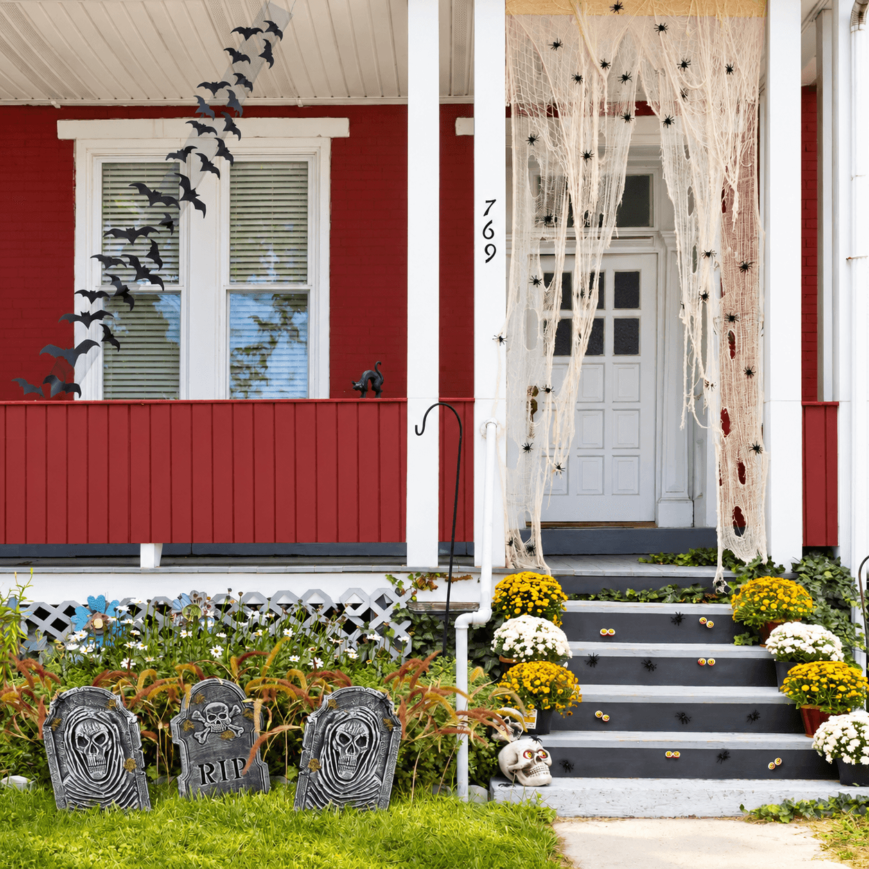 Red front porch decorated for Halloween with hanging black bats, white faux spiderwebs covered in black spiders, gravestones in the garden, pumpkins and flowers along the steps, and a skull decoration near the walkway.