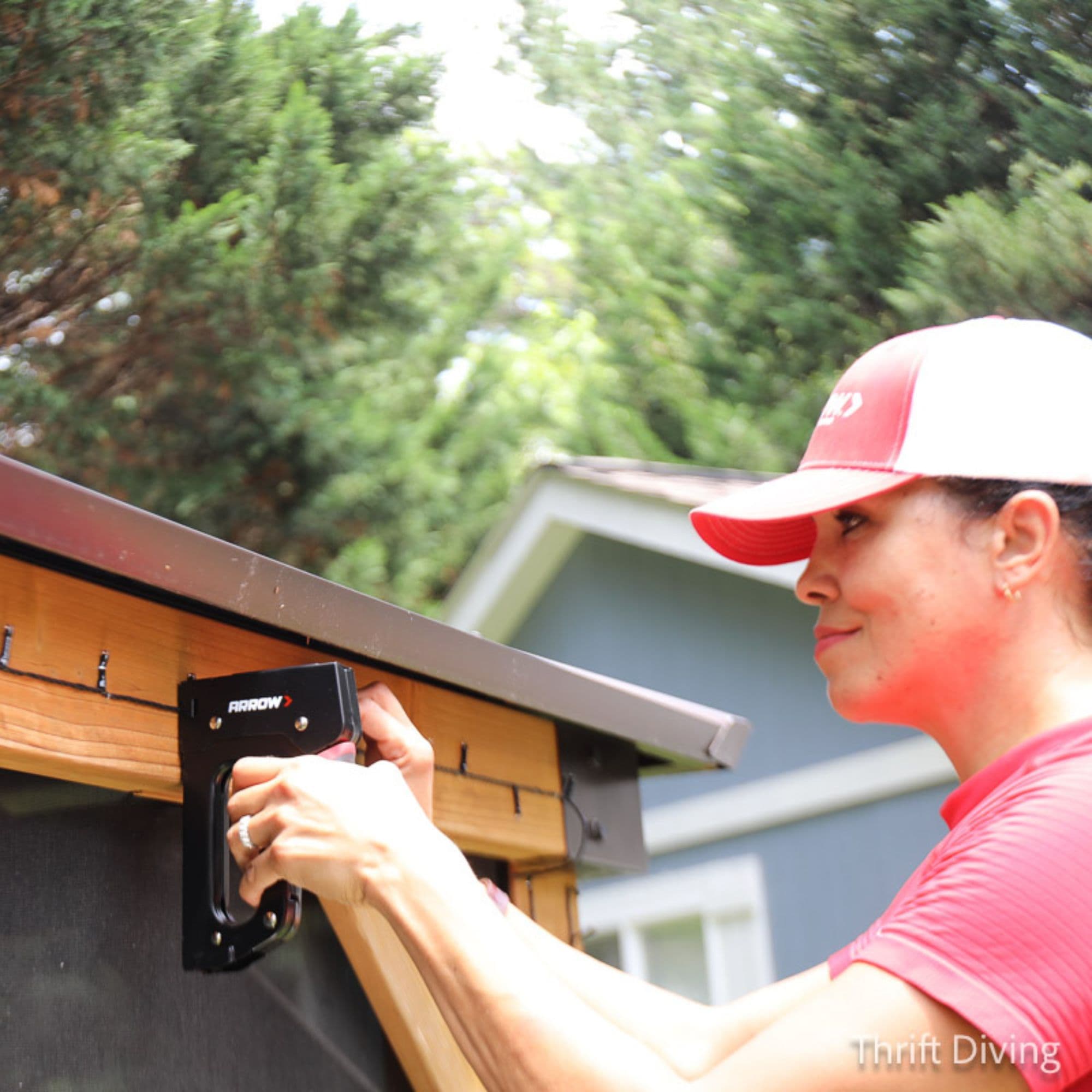 Serena, a woman in a red hat, staples string lights to the wooden top edge of her gazebo using a staple gun.