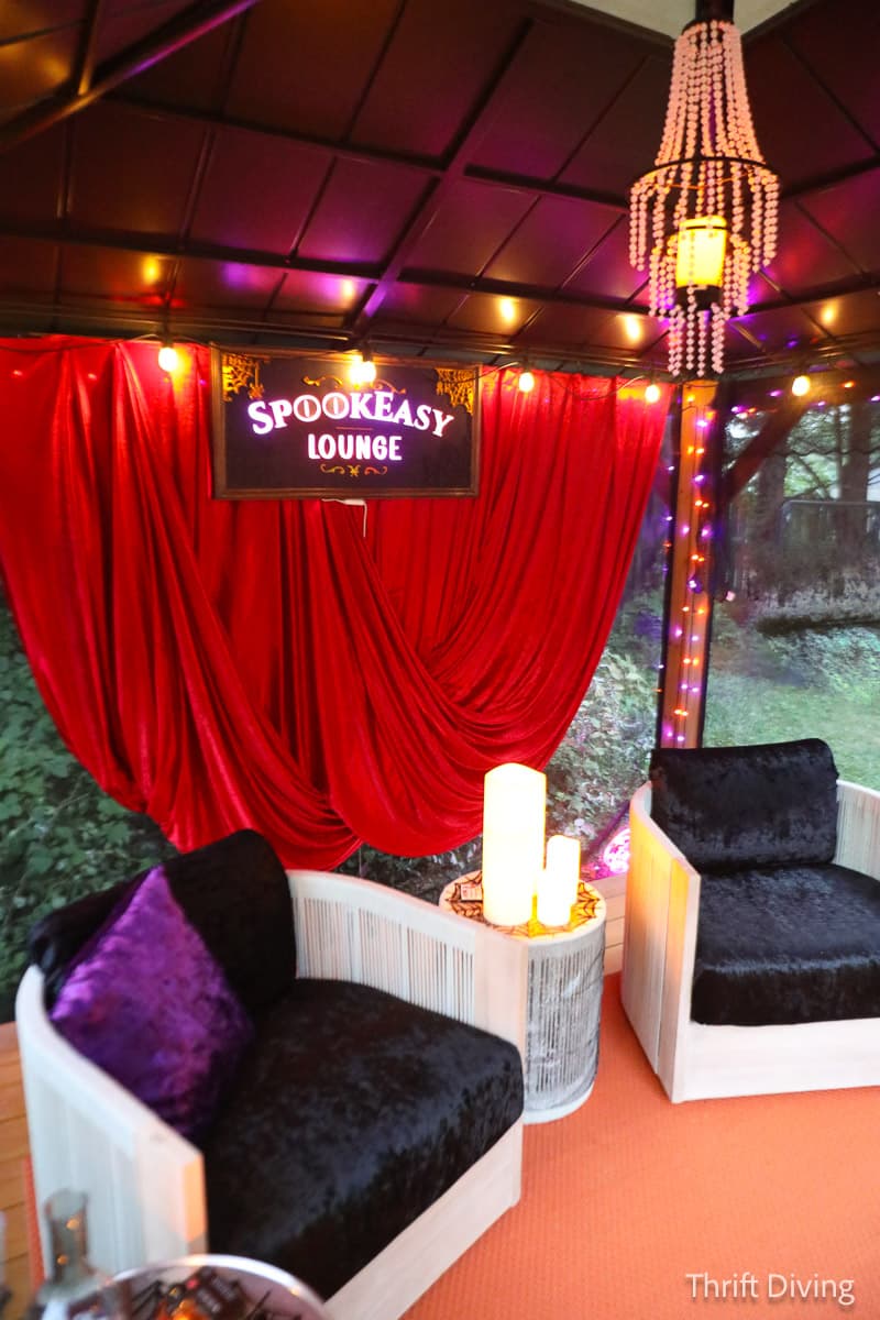 The interior of the Spookeasy Lounge, featuring two plush chairs, a red curtain, LED candles, a purple ceiling tarp, and a beaded chandelier.