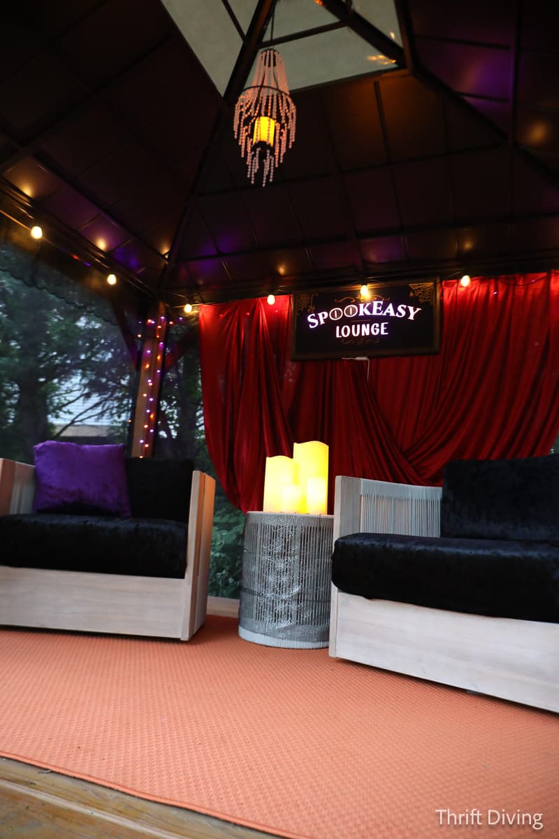 The interior of the Spookeasy Lounge, featuring two plush chairs, a red curtain, LED candles, a purple ceiling tarp, and a beaded chandelier.
