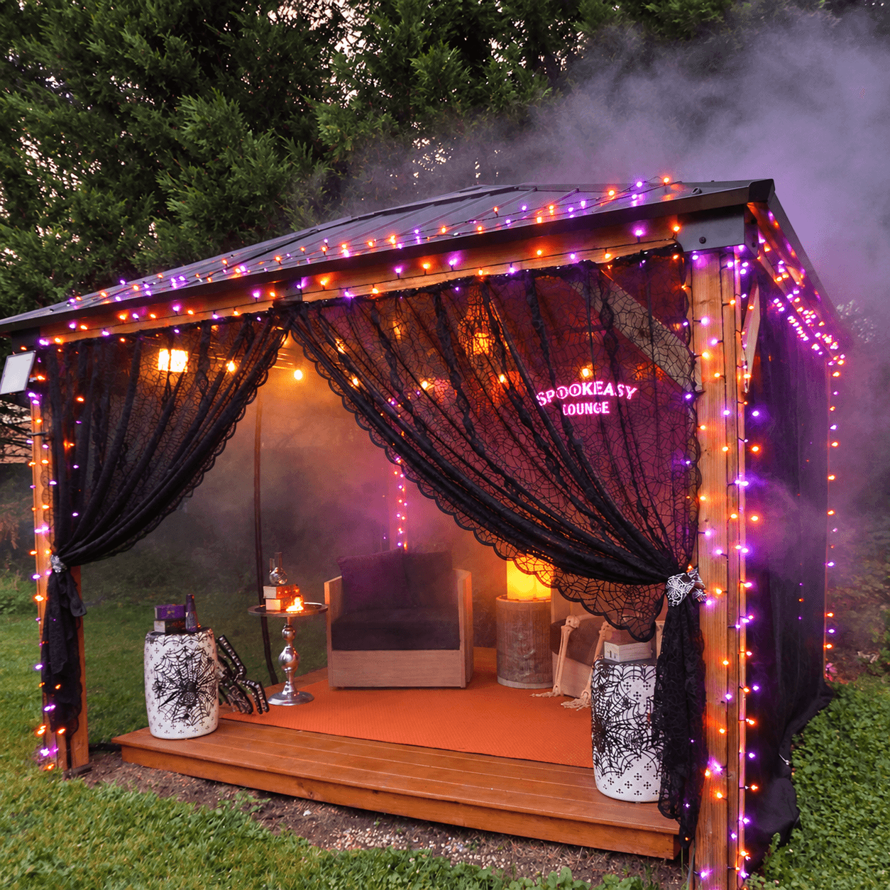 Outdoor Halloween gazebo decorated with black spiderweb curtains, purple and orange string lights, and a glowing Spookeasy Lounge sign.