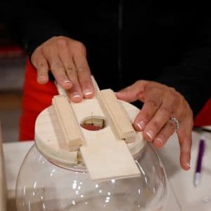 Person sliding a rectangular wooden piece between two guide rails on a circular wooden lid atop a clear glass bowl for a DIY candy dispenser.