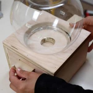 Person positioning a clear glass bowl on top of an unfinished plywood candy dispenser, aligning it with the circular center opening.