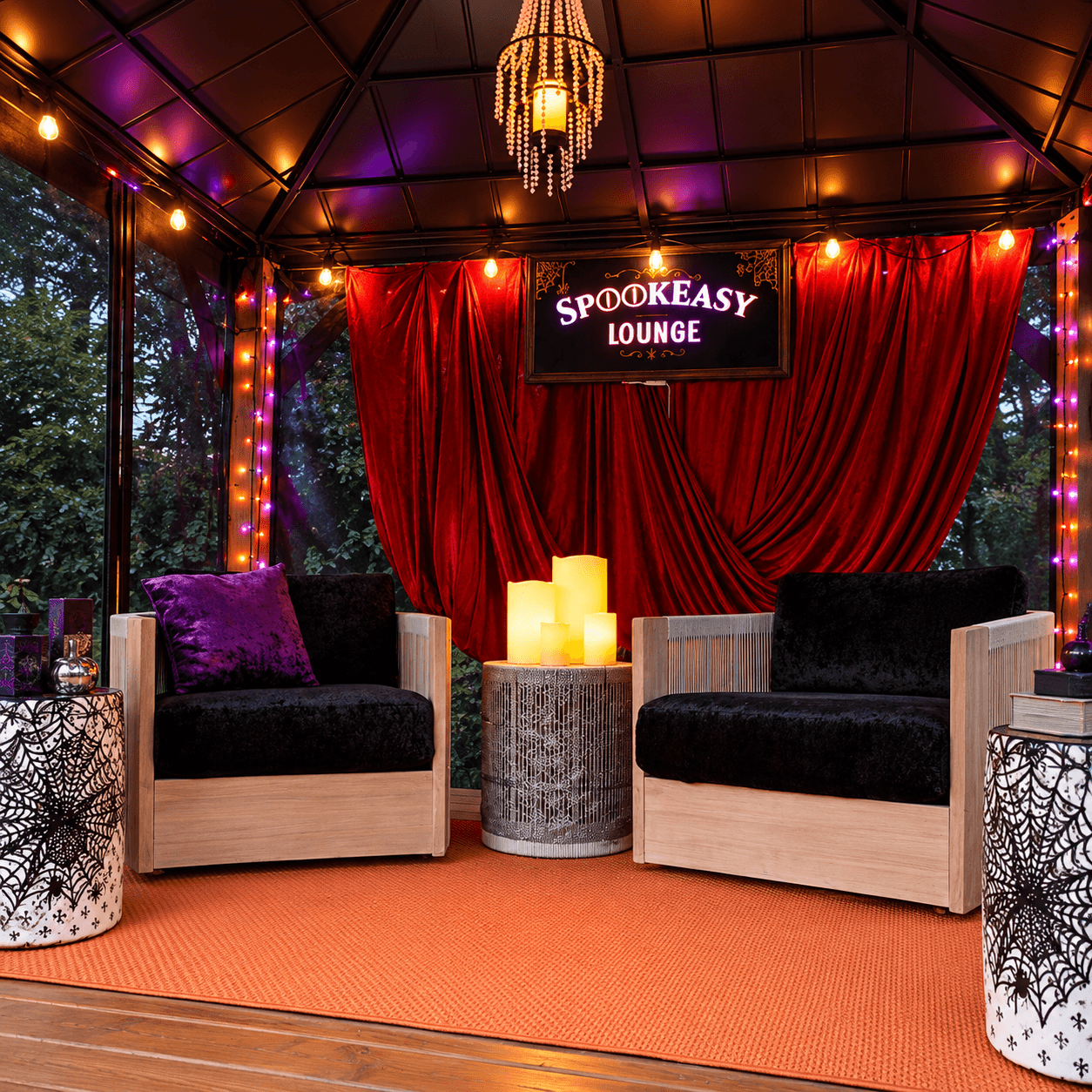 Cozy Halloween lounge inside a backyard gazebo with black cushioned chairs, red velvet curtains, glowing candles, and spiderweb decor.