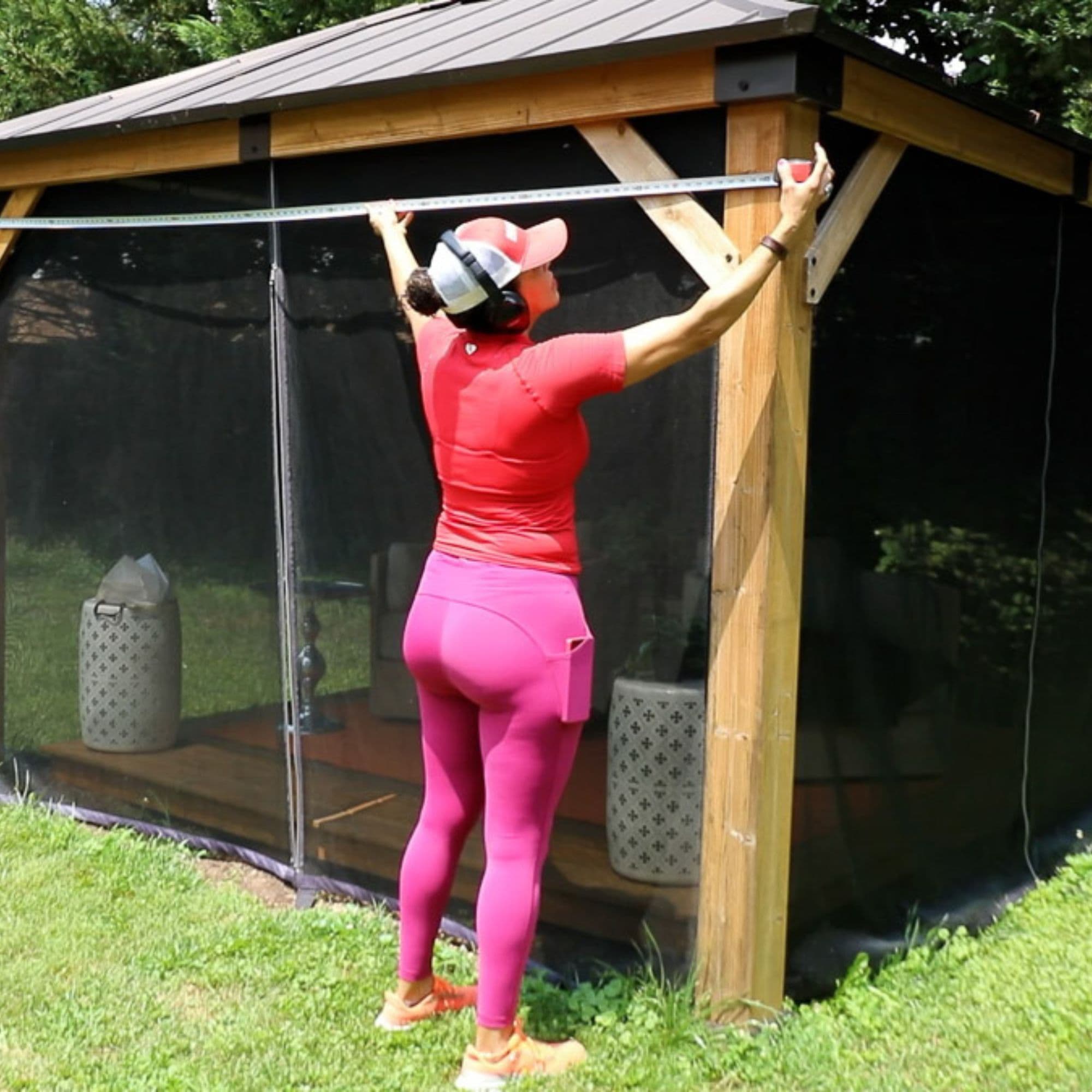 Serena, a woman wearing a red hat, earphones, red t shirt, and pink leggings with orange sneakers, is measuring the width of her gazebo.