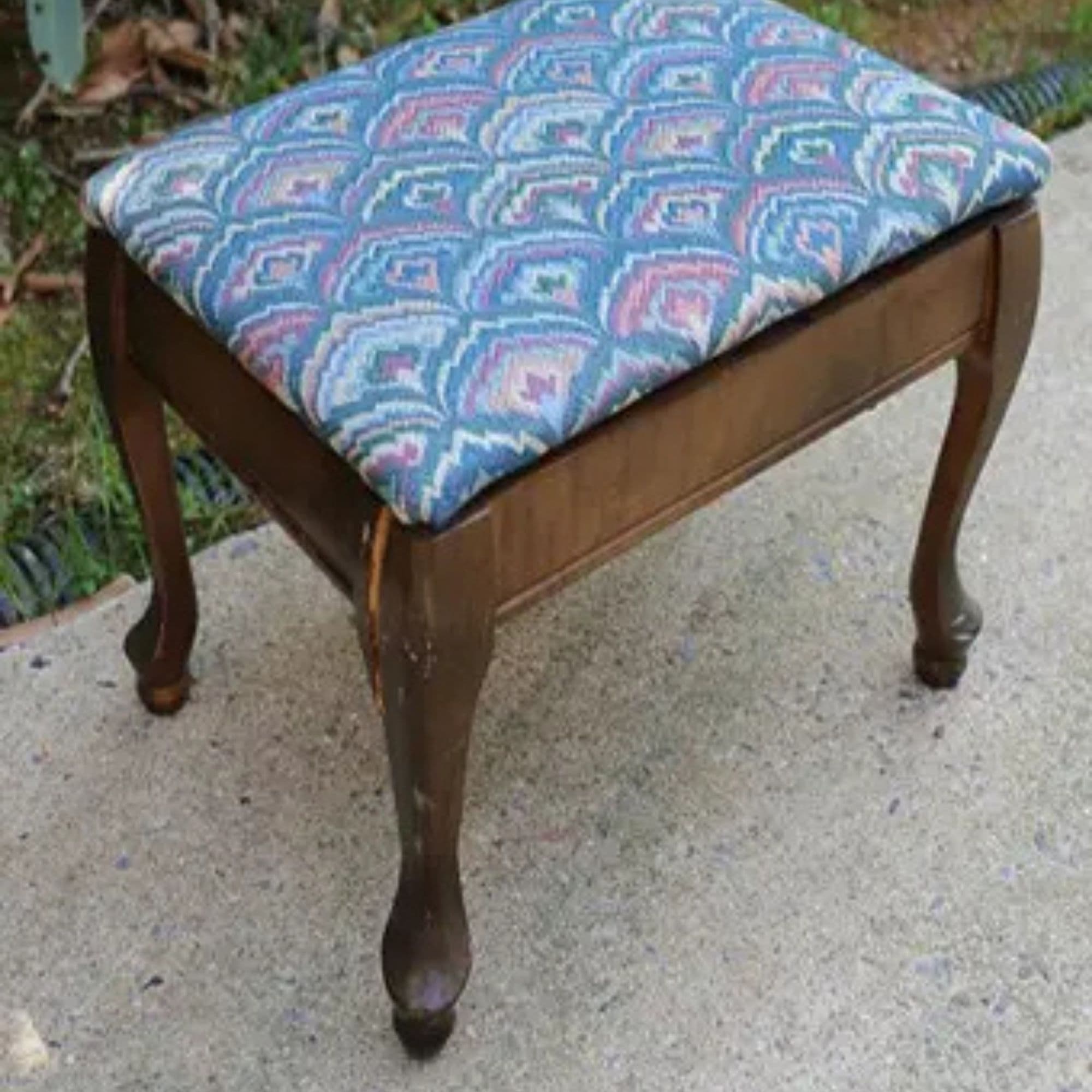 Vintage wooden stool with curved legs and a blue-and-pink patterned upholstered cushion on a concrete patio.