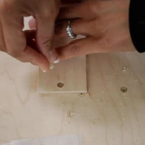 Person inserting a small wooden dowel into a drilled hole in a rectangular wood piece during assembly of a DIY candy dispenser.