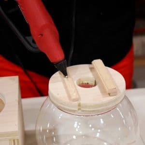 Person using a red hot glue gun to attach wooden strips to a circular wooden lid on top of a clear glass bowl for a DIY candy dispenser.
